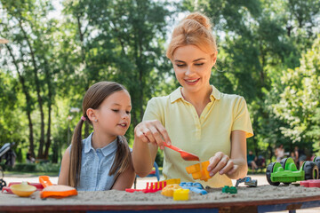 smiling woman playing with toy shovel and sand near daughter.