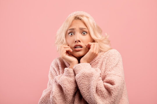 Young caucasian woman feeling scared, touching cheeks with hands, posing on pink studio background