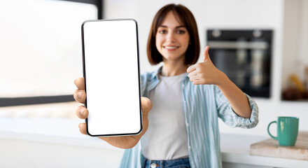 Online offer. Happy lady standing in kitchen, demonstrating smartphone with blank screen and showing thumb up