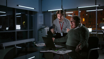 Happy colleagues look at laptop screen and celebrate success working together late in dark office