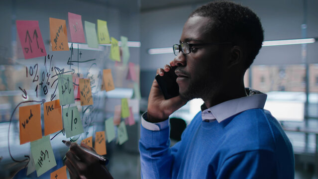 Side view of African-American businessman in mask write on transpaent board talking on cellphone - Powered by Adobe