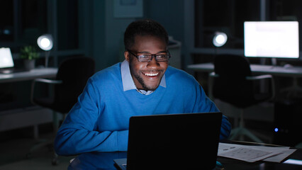 Portrait of happy African-American businessman look at laptop screen and smile in dark empty office