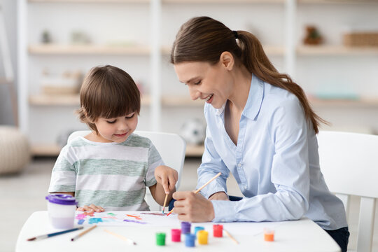 Cute little boy enjoying art classes, drawing picture with colorful paints, sitting at desk with teacher and laughing