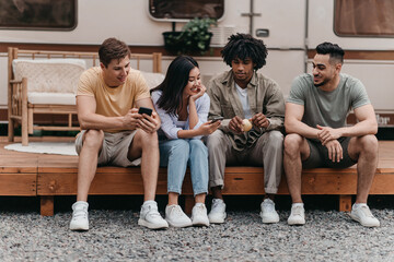 Group of diverse friends sitting near camper van, speaking to each other, using smartphones on camping trip