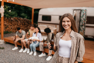 Smiling young woman holding bottle of beer, having fun camping trip together with her multiracial friends outdoors