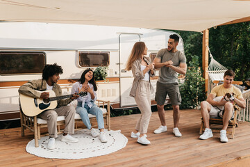 Young diverse friends with dog having fun near camper van, playing guitar, singing and dancing, enjoying summer vacation - Powered by Adobe