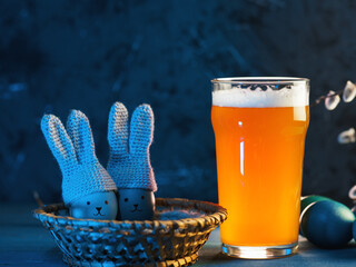 Easter eggs in crocheted hats with bunny ears and a glass of craft beer. Blue background, holiday...