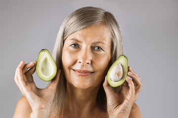 Portrait of pleasant mature woman posing in studio with halves of green ripe avocado in hands. Caucasian lady recommending organic vitamin cosmetic for beauty routine.