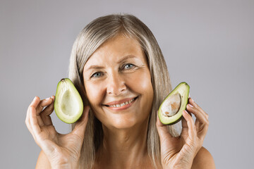 Portrait of pleasant mature woman posing in studio with halves of green ripe avocado in hands. Caucasian lady recommending organic vitamin cosmetic for beauty routine.