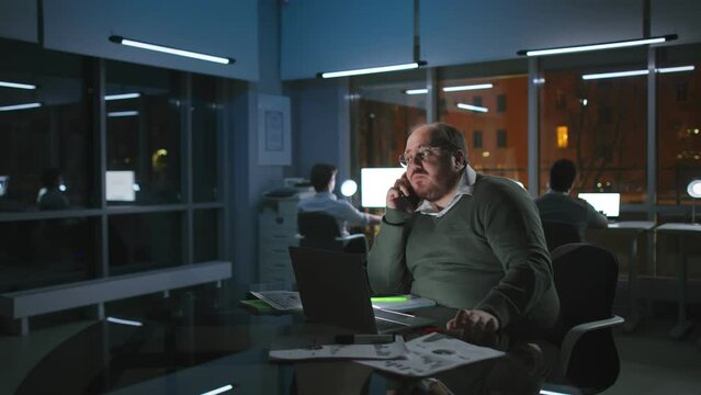 Fat call center worker talk on cellphone with client working late in dark office