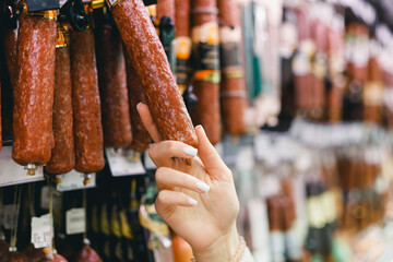Woman chooses dried smoked sausage hanging in refrigerator in butcher's shop