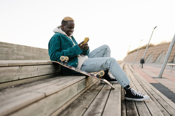 Happy young african man with skateboard looking at mobile phone social media app to connect with...