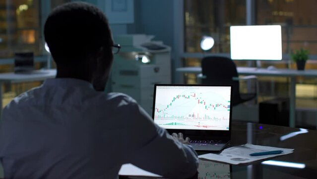 Back view of African-American businessman work with graphs on laptop late in empty office.