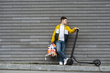 Modern teenager with a backpack and electric scooter stands near a brick wall