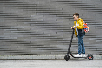 Boy with a backpack rides on electric scooter against a gray brick wall © Andrii 