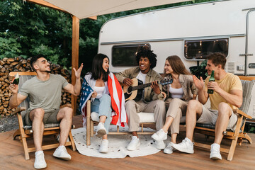 Interracial friends playing guitar and singing, holding American flag, drinking beer, sitting near...