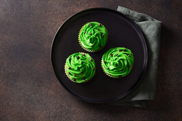 Three chocolate cupcakes with green whipped cream and chocolate sprinkles on brown background. St....