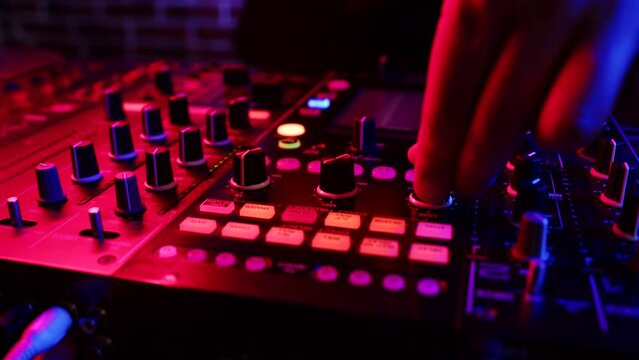 DJ twists knobs on mixing console behind his counter to set up rave in nightclub, closeup. DJ works in disco club lit by neon