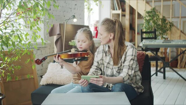 A woman helps a girl to play the violin. She watches notes on her phone