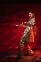 Fototapeta premium Fashionable woman wearing sequin suit, posing, dancing against red background. Full-length studio fashion portrait with move effect and art lighting 