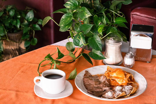 Chilaquiles con bistec, huevo estrellado y frijoles, acompañados de taza de café, planta teléfono y azucarero en restaurante. 