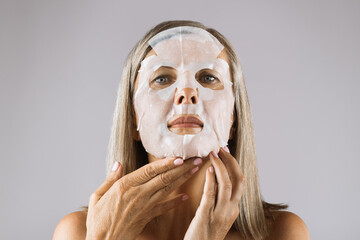 Beautiful senior woman applying white fabric mask on face over grey studio background. Caucasian...