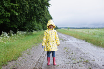 Happy child in a yellow raincoat walks in the rain on a country road