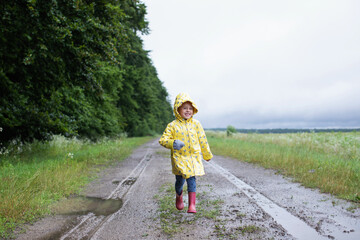 Little girl in a yellow raincoat runs on a dirt road with puddles and mud