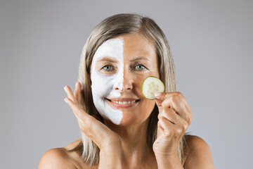 Portrait of smiling aged woman holding slice of cucumber with while clay mask on half of face. Caucasian female with bare shoulders isolated over grey background.