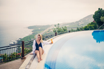 Beautiful attractive young woman sits near a large pool overlooking the sea. Vacation concept. Girl in a black swimsuit smears her skin with sunscreen