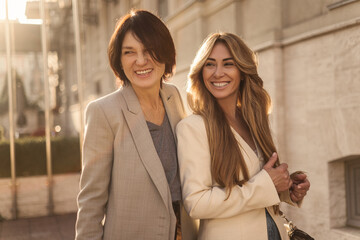 Two caucasian adult women brunette and blonde are laughing looking into distance while standing outdoors. Ladies in trendy casual clothes enjoy walk together. Happy weekend concept