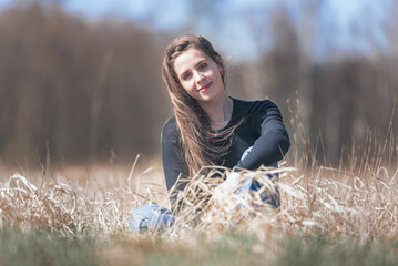 Naklejka na ściany i meble Young lady in casual clothes sitting on the grass. The girl enjoys the rays of the sun that fall on her. The grass is dry and there is a forest in the background. It is a sunny day.