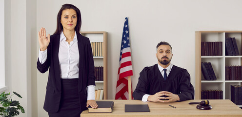 American witness takes an oath before giving a testimony. A jury member standing near a male judge in the court room swears on the Bible that she will tell the truth. Law and justice in the US concept - obrazy, fototapety, plakaty