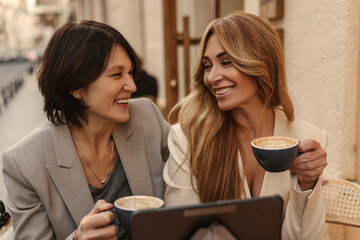 Happy caucasian adult women laugh, look at each other while sitting in cafe with coffee and tablet. Ladies at lunch are celebrating deal at work. Positive human reactions and emotions