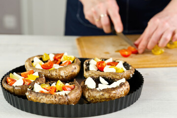 raw champignons stuffed with cheese, tomatoes and paprika in a baking dish. Woman hands cuts tomatos on the background. close up