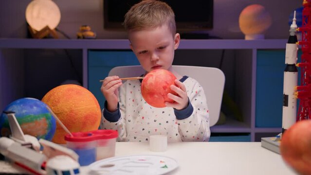 Blond boy paint planet solar system Mercury with colorful paint sitting home table in evening, planet solar system, spaceships and space shuttle from constructor around. Cosmonautics Day on April 12.