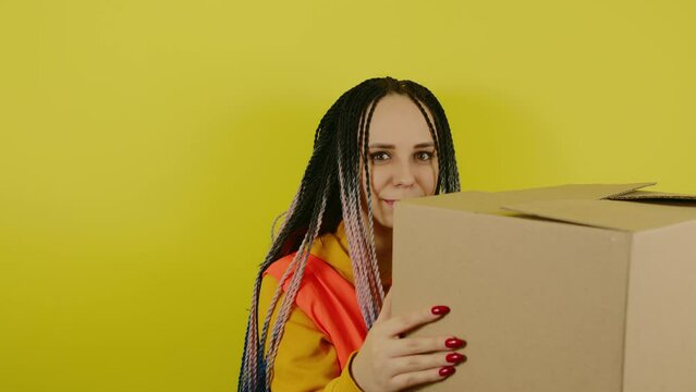 Young woman in vest with cardboard box on yellow background in studio. Female courier with package. Concept of delivery and transportation.