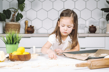 Portrait of little studiously girl chef, sitting on chair, making different shape dough cookies on...