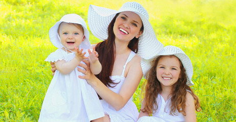 Portrait of happy smiling mother with two daughters children wearing summer straw hats on the grass in park