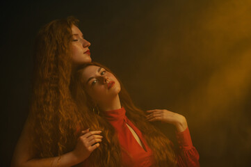 Studio art portrait of two beautiful redhead freckled women with long natural curly hair, red lips makeup posing in darkness and warm light. Copy, empty space for text
