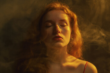 Beautiful redhead freckled woman posing in darkness, smoke and warm light. Art studio portrait....