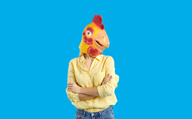 Person in yellow shirt and chicken mask on head standing with arms folded, isolated on blue studio...