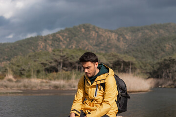 Young handsome man sitting on old small dock. He is looking his reflection on water. He is sad. Loneliness. Wearing yellow coat. Beautiful landscape.