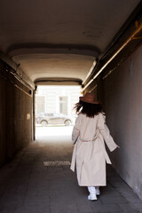  curly brunette girl in sun glasses and hat going away outdoors. Young woman happy walking in street. Pink sweater, beige coat, beige hat. Sun in city. Fashionable asian girl with frizzly hair