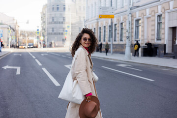 Fototapeta premium Happy curly brunette girl in sun glasses smiling outdoors. Young woman happy walking in street. Pink sweater, beige coat, beige hat. Sun in city. Fashionable asian girl with frizzly hair