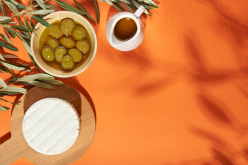 Set of green olives with olive oil and white cheese on orange background. Spring sunlight with soft shadows. Healthy Mediterranean food. Top view, flat lay, copy space