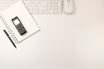 White keyboard, mouse and notepad on left side with black pen and voice recorder flat lay on white background