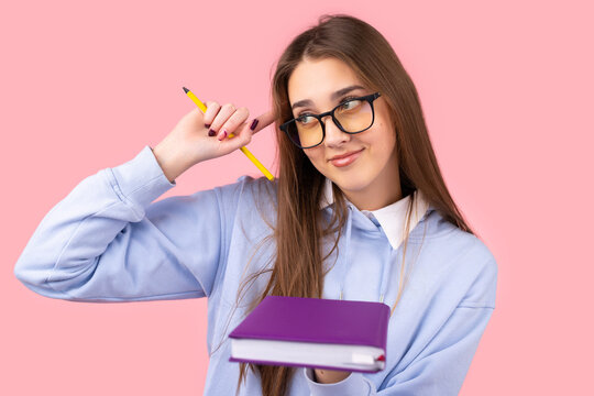 Jiyful student girl with long blond hair in glasses holding a book in one hand and keeping a pencil in another holding an index finget on the temple.