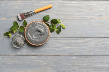 Bowls of activated carbon facial mask, powder, makeup brush and plant branches on grey wooden background
