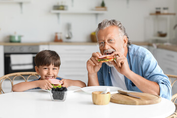 Little boy with his grandfather eating tasty sandwiches at table in kitchen